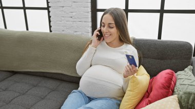 Telefonda konuşurken gülümseyen ve evde kredi kartı tutan hamile İspanyol kadın.
