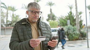 Akıllı telefon ve kredi kartını şehir yaşamını ve teknolojiyi gösteren bir sokakta ödeme yapmak için kullanan olgun bir adam..