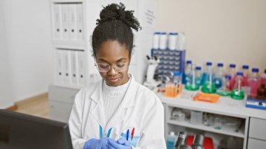 Laboratuvar ortamında kimyasal mataralar ve ekipmanlarla örnekleri analiz eden laboratuvar önlüklü Afrikalı Amerikalı kadın bilim adamı..