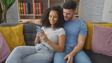 Bir adam ve bir kadın, rahat bir oturma odasında akıllı telefona bakarken sarı ve pembe yastıklı gri bir kanepede sarılıyorlar..