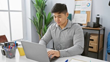 Modern ofis ortamında dizüstü bilgisayarla çalışan odaklanmış genç Asyalı adam üretkenlik ve profesyonelliği aktarıyor..