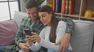 İspanyol çift kapalı koltukta rahat, akıllı telefona gülümsüyor, rahat bir evde sevgi ve beraberlik sergiliyor..