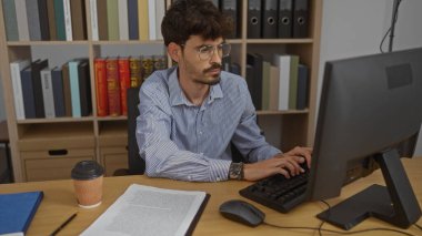 Ofiste çalışan sakallı genç bir adam, klavyede daktilo kullanıyor, kitaplarla ve dosyalarla çevrili, gözlük takıyor, ekrana odaklanmış, ön planda kahve fincanı var..