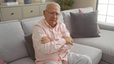 Oturma odasında oturan, kolları çaprazlanmış, pembe bir gömlek ve gözlük takmış, rahat mobilyalar ve hafif yastıklarla çevrili, düşünceli beyaz bir adam..