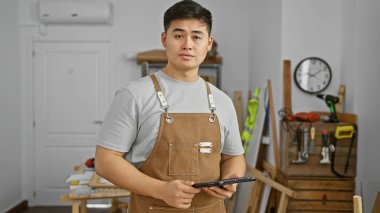 Kahverengi önlüklü Asyalı adam, elinde aletlerle marangozluk stüdyosunda tablet tutuyor.