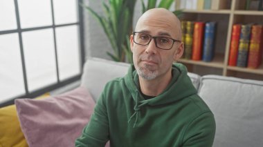 Gözlüklü, yeşil kapüşonlu, sakallı yakışıklı, kitaplık arka planda, kapalı koltukta oturan kel adam.