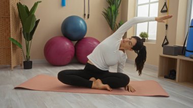 Spor salonunda egzersiz topları, minder, bitkiler ve spor aletleriyle yoga yapan genç bir kadın. Parlak bir odada esniyor ve rahat görünüyor.