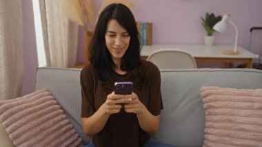 Yumuşak yastıkları ve pastel dekoru olan rahat bir apartman dairesinde akıllı telefon kullanan bir kadın gülümsüyor..