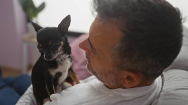 Orta yaşlı İspanyol bir adam sıcak bir oturma odasında rahatlarken Chihuahua 'sını sevgiyle tutuyor..