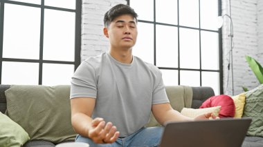 Genç Asyalı bir adam modern bir oturma odasında meditasyon yapıyor. Huzur ve sükunet yayıyor..