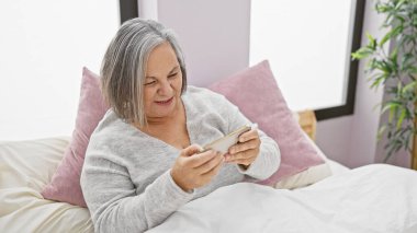 Gülümseyen, son sınıf öğrencisi bir kadın rahat bir yatak odası ortamında akıllı bir telefonla konuşurken yatakta uzanıyor..