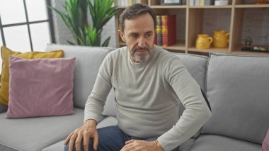 Orta yaşlı, sakallı bir adam, rahat, modern bir oturma odasında, gri bir koltukta düşünceli bir şekilde oturuyor..