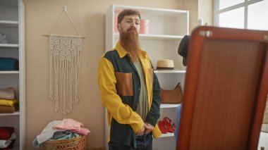 Renkli ceketli kızıl saçlı adam kumaş ve kıyafetlerle terzi dükkanında poz veriyor..