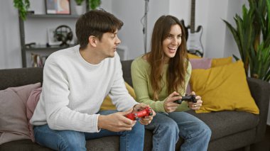 Bir erkek ve kadın, bitki dolu modern bir oturma odasındaki rahat bir kanepede video oyunu oynamaktan zevk alırlar..