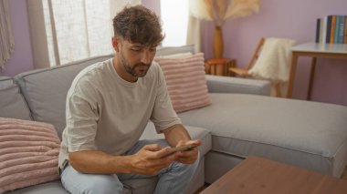 Genç adam rahat bir oturma odasında kanepede oturuyor, akıllı telefonuna düşünceli bir ifadeyle odaklanıyor..