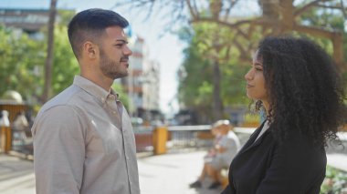 Gülümseyen bir çift yeşillik içinde güneşli bir caddede yan yana durur..