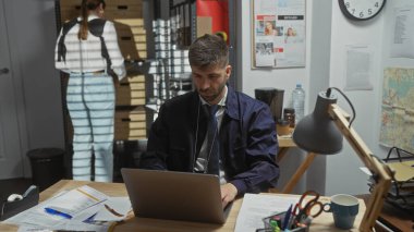 Man and woman working as a team in a well-equipped indoor police station, with the man focused on a laptop. clipart