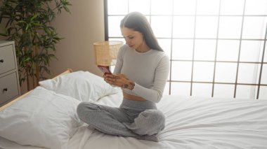 Evdeki parlak bir yatak odasındaki yatakta oturan genç bir kadın telefonunu kullanıyor. Rahatlama ve modern yaşam tarzını evde aktarıyor..