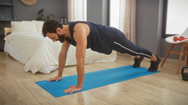 Sakallı bir adam modern bir yatak odası ortamında mavi bir paspas üzerinde tahta egzersizi yapıyor.