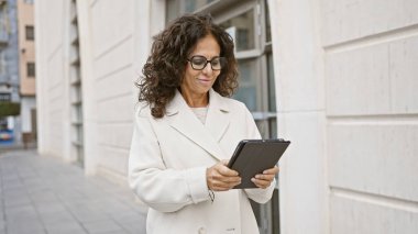 Şehir caddesinde tablet kullanan olgun İspanyol bir kadın, gelişigüzel şehir işlerini somutlaştırıyor..