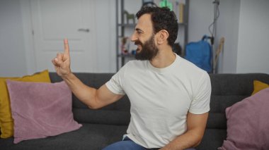 Oturma odasındaki neşeli sakallı bir adam yukarı bakıyor, sevinç gösterisi yapıyor ya da bir fikri var..