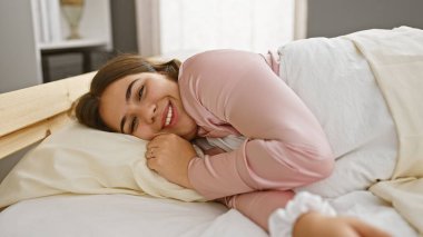 Gülümseyen genç bir İspanyol kadın rahatça bir yatak odasında uzanıyor, rahat, samimi ve çekici bir hava yayıyor..