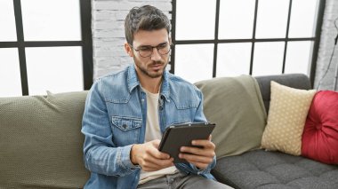 Sakallı genç bir İspanyol, modern bir oturma odasındaki kanepede otururken tablet kullanıyor.