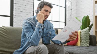 Odaklanmış İspanyol bir adam modern bir oturma odasında telefonla konuşurken bir belgeyi analiz eder..
