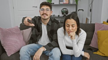 Sıkılmış bir kadın ve nişanlı bir adam modern bir oturma odasında uzaktan kumandayla birlikte koltukta oturuyorlar..