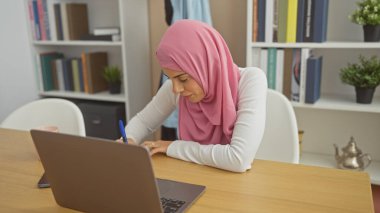 Başörtüsü takan genç bir İspanyol kadın evdeki dizüstü bilgisayarında çalışırken not alıyor ve eğitim rutinine göz atıyor..