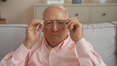 Yaşlı, beyaz bir adam oturma odasında otururken gözlüğünü ayarlıyor. Yetişkinliğini evin içinde sergiliyor..