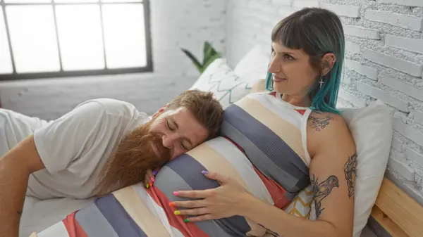 stock image Couple enjoying a tender moment as man listens to baby's movements by resting his head on pregnant woman's belly while lying on a bed in a cozy bedroom.