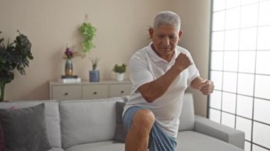 Olgun bir İspanyol adam modern, iyi dekore edilmiş bir oturma odasında egzersiz yapıyor. Evinde bitki ve rahat mobilyalarla sağlığını ve canlılığını somutlaştırıyor..