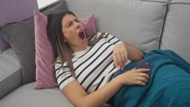 Genç, ispanyol, esmer bir kadın oturma odasındaki bir kanepede uyur, akıllı telefonunu bir deniz mavisi battaniyesinin altında tutar, rahat bir ev dekorasyonu sergiler..