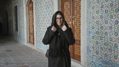 İstanbul 'daki Topkapı Sarayı' nda süslü döşemelerin önünde duran gözlüklü ve eşarplı gülümseyen genç kadın, hindi.