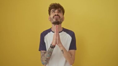 Duygusal genç İspanyol bir adam izole edilmiş sarı bir arka plana karşı tek başına duruyor elleri dua ederken umut dolu bir ifadeyle yalvarıyor.