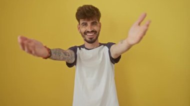 Neşeli genç İspanyol adam kollarını açmış, mutluluğu kucaklıyor ve sevgiyi kucaklıyor; neşeli sarı bir arka planda izole edilmiş, kameraya gülümsüyor.