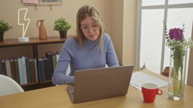 İki kadın evlerinde etkileşim içinde, biri dizüstü bilgisayarına yoğunlaşırken diğeri yanında duruyor, rahat bir apartman dairesinde..