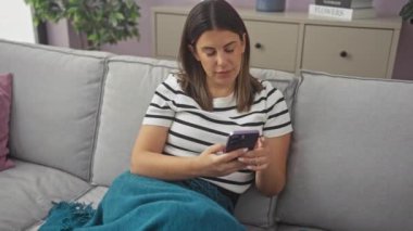Genç bir İspanyol kadın rahat bir oturma odasında, battaniyeye sarılmış, rahat bir kanepede dinlenirken akıllı telefonunu kullanıyor..