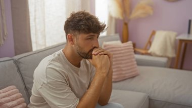 Düşünceli bir genç adam, rahat bir oturma odasında pastel dekorlu ve yumuşak ışıklandırmalı rahat bir kanepede, düşünceli bir ifadeyle evin içini yansıtıyor..