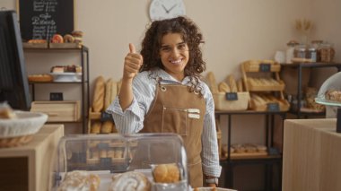 Young woman in a bakery giving a thumbs-up while surrounded by freshly baked bread and pastries indoors during the daytime clipart