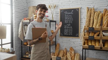 Pastanede sakalı ve panosu olan genç İspanyol adam beyaz tuğla duvara karşı arkaplanda hamur işleri ve ekmeklerle kara tahta menüsünü işaret ediyor.