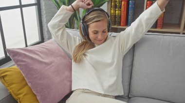 Kafkasyalı genç bir kadın, rahat bir oturma odasında, kanepede dans etmekten ve kulaklıkla müzik dinlemekten zevk alır..