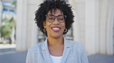 Kıvırcık saçlı ve gözlüklü gülümseyen Afro-Amerikalı kadın şehir ortamında açık havada duruyor, açık renk ceket ve geniş halka küpeler takıyor, şehir binaları arka planda bulanıklaşıyor..