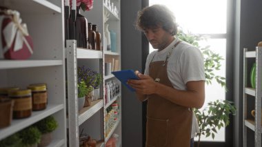 Önlüklü genç adam, çeşitli dekorasyon ürünleri ve bitkilerle çevrili bir dekor dükkanında tablet kullanıyor.