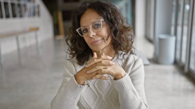 Kendinden emin, orta yaşlı İspanyol bir kadın, iyi aydınlatılmış bir ofis içinde, profesyonellik ve deneyim dolu bir şekilde poz veriyor..