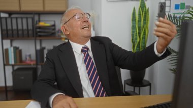 Takım elbiseli, gri saçlı yaşlı bir adam arka planda raflar ve bitkiler olan bir ofis ortamında gülüyor ve selfie çekiyor..
