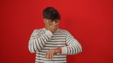 Genç bir İspanyol erkeği, eli ağzında, izole edilmiş kırmızı bir arka planda şaşkın şaşkın bakar..