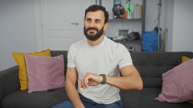 Gündelik beyaz gömlekli sakallı yakışıklı bir adam renkli yastıklarla modern bir oturma odasında oturuyor..