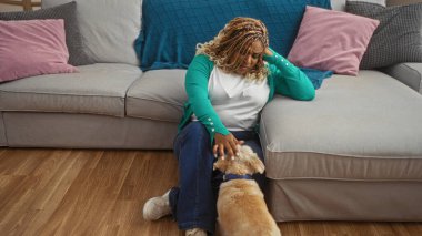 Saçları örgülü genç bir Afrikalı Amerikalı kadın oturma odasının zeminine oturmuş renkli yastıkları olan bir köpeği şefkatle okşuyor..
